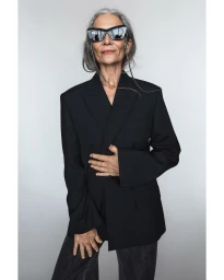 Elderly woman with long grey hair wearing oversized cat-eye sunglasses and a black blazer against a light grey background.