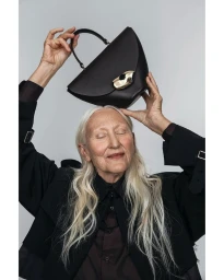 Elderly woman with long silver hair holding a structured black handbag above her head against a grey seamless background