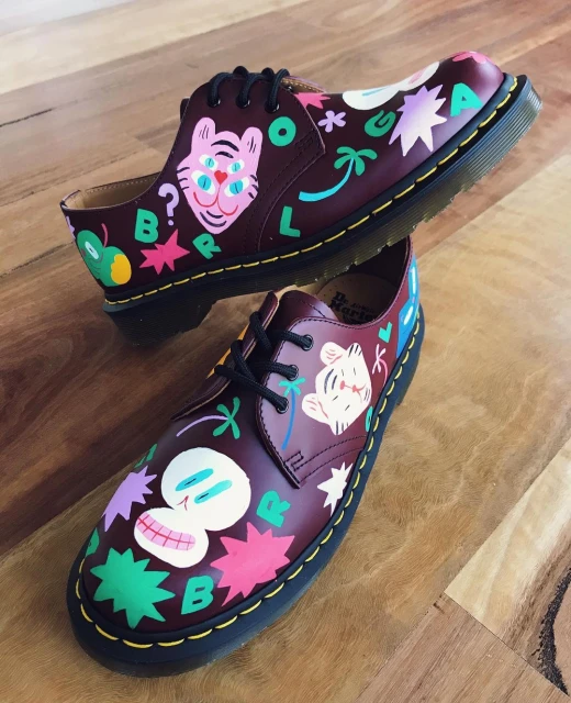 Pair of burgundy Dr. Martens 1461 shoes with colorful hand-painted designs, including skulls, tigers, stars, and letter shapes, on a wood surface.