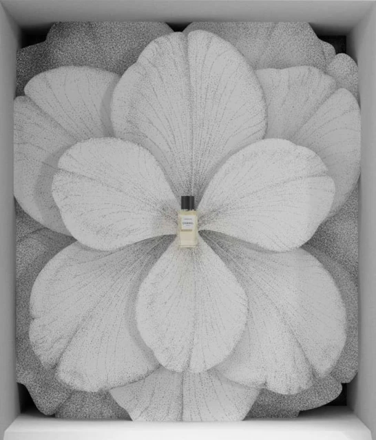 Close-up of a small perfume bottle centered against a large flower backdrop inside a white display case, product-focused composition.