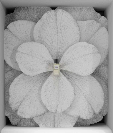 Close-up of a small perfume bottle centered against a large flower backdrop inside a white display case, product-focused composition.