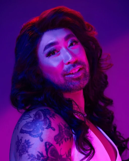 Headshot of a person with facial hair and long wavy hair, wearing a white top, posing against a purple and blue background.