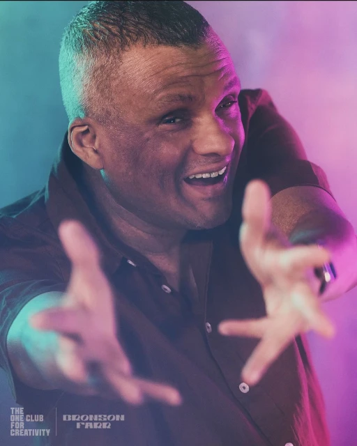 Adult man in a button-down shirt extends his hands out against a backdrop of colored lights and smoke in an indoor environment.