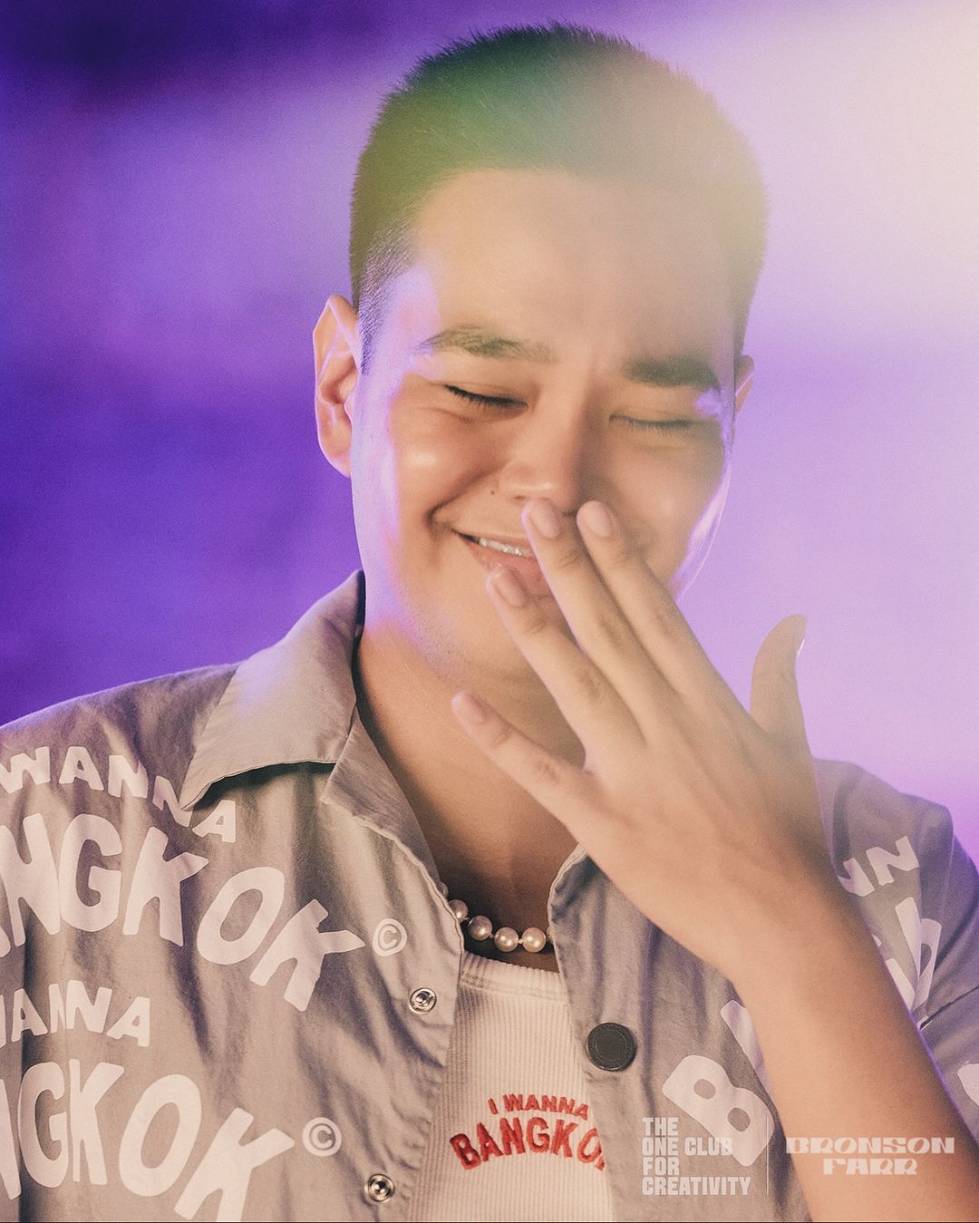 Close-up of a smiling Asian man with a buzzcut, wearing a Bangkok-themed shirt, with a hand near his face against a purple backdrop.