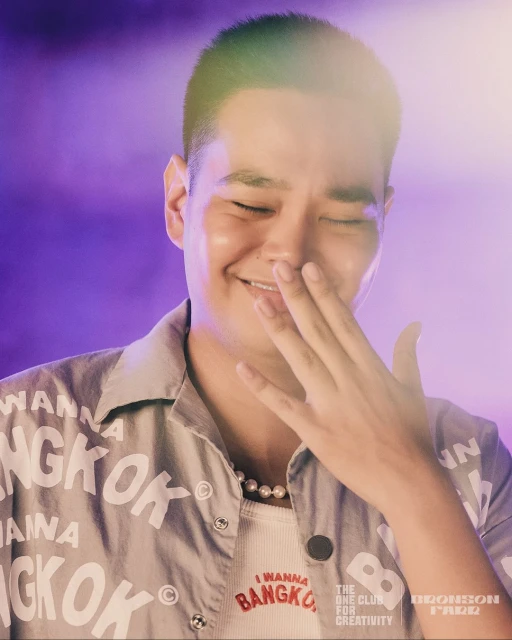 Close-up of a smiling Asian man with a buzzcut, wearing a Bangkok-themed shirt, with a hand near his face against a purple backdrop.