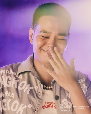 Close-up of a smiling Asian man with a buzzcut, wearing a Bangkok-themed shirt, with a hand near his face against a purple backdrop.