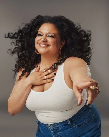 Smiling woman with long dark curly hair wearing a white tank top and blue jeans pointing toward the camera