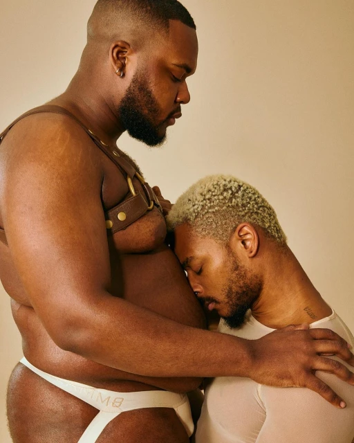 Two adult Black men pose closely against a light background. One man wears a brown leather harness and a white jockstrap.