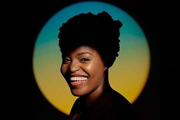 Portrait of a smiling Black woman with dark, afro hair against a backdrop of a glowing, diffused circle of blue and yellow light.