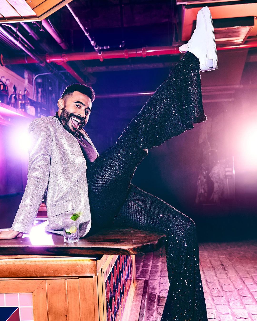 A man poses on a bar in a sequined jacket and pants, with one leg raised, cocktail visible, showcasing fashion and nightlife.