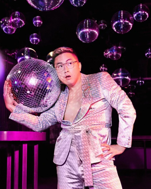 An Asian man in a patterned suit leans against a disco ball in a club, surrounded by hanging disco balls and neon purple light.
