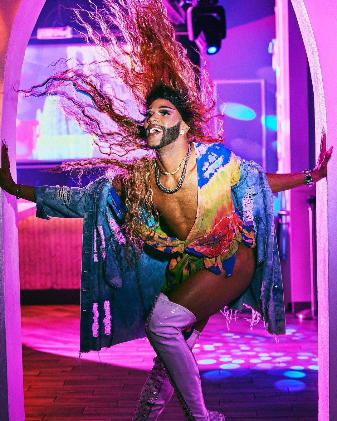 A person in drag poses in a doorway with dramatic hair, wearing a colorful shirt and jean jacket, and light purple knee-high boots.