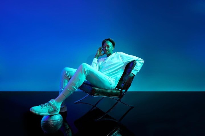 Woman poses in athletic wear, seated on chair with a basketball under her foot, against blue backdrop in studio lighting.