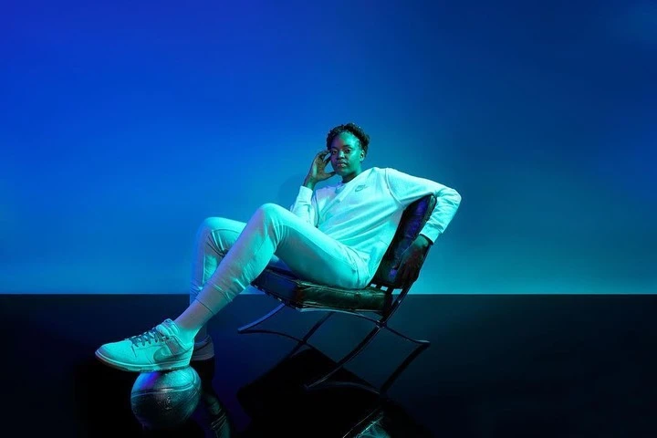 Woman poses in athletic wear, seated on chair with a basketball under her foot, against blue backdrop in studio lighting.