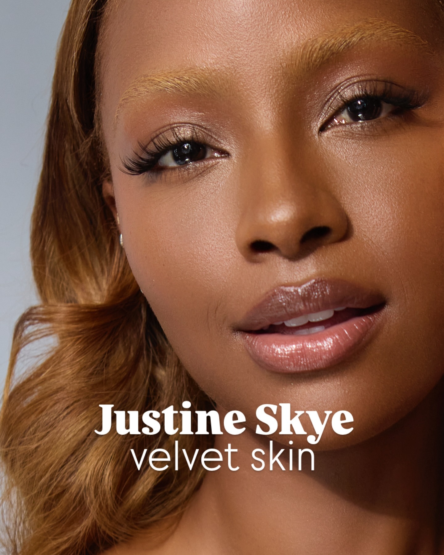 Close-up portrait of a brown-skinned woman with honey-blonde hair and glossy lips, in soft studio lighting.