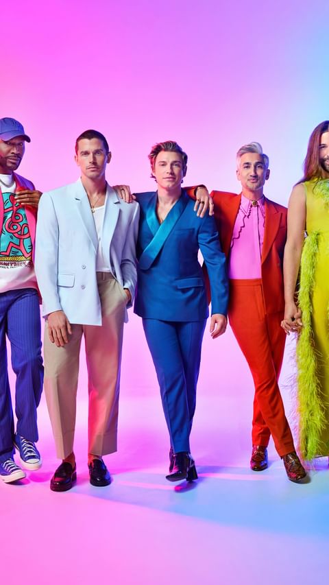 Promotional shot of five stylish cast members standing together, wearing bright colorful suits and outfits, on a graduated pink and blue background.