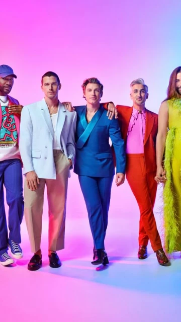 Promotional shot of five stylish cast members standing together, wearing bright colorful suits and outfits, on a graduated pink and blue background.