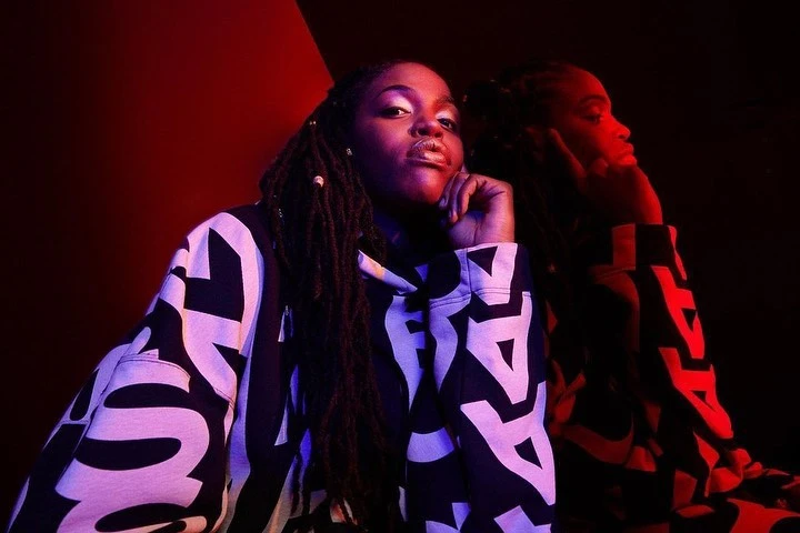 Portrait of a Black woman with dreadlocks wearing a patterned hoodie, gazing at the camera with a reflective red image in the background.
