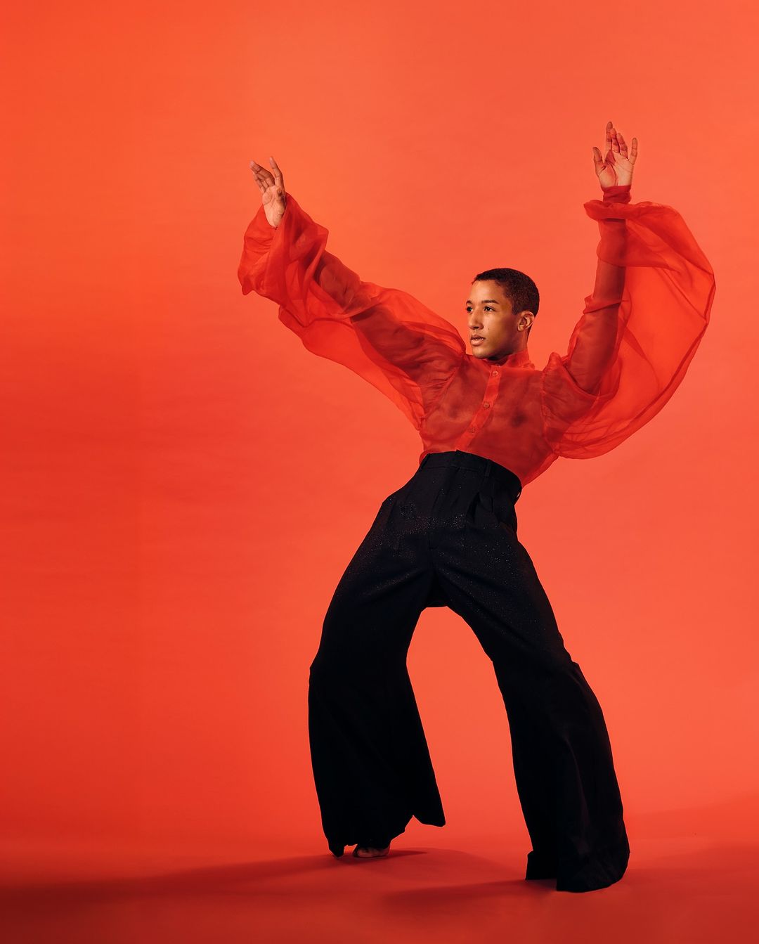 Young adult male in wide-legged black pants and sheer red top posing on red studio background with arms raised in modern dance fashion pose.