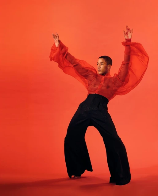 Young adult male in wide-legged black pants and sheer red top posing on red studio background with arms raised in modern dance fashion pose.