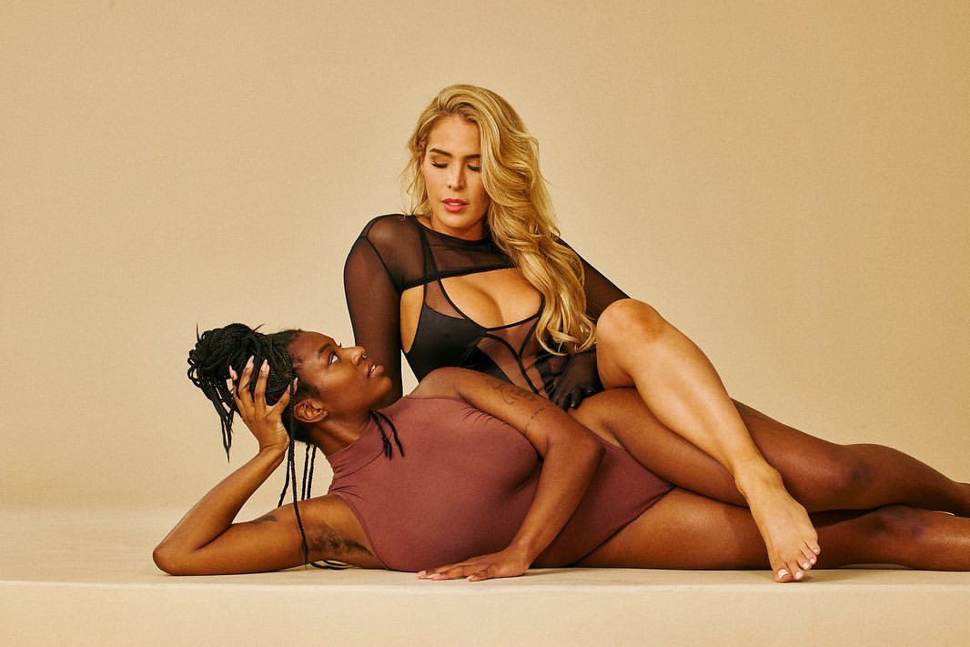 Two women in bodysuits pose against a beige backdrop. One woman with long blonde hair rests over the other with braided black hair.