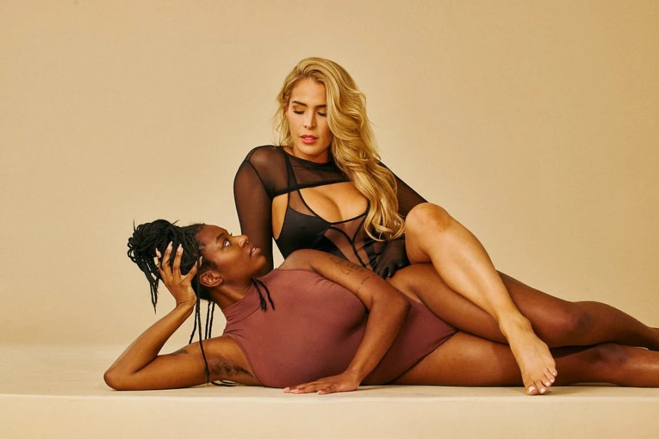 Two women in bodysuits pose against a beige backdrop. One woman with long blonde hair rests over the other with braided black hair.