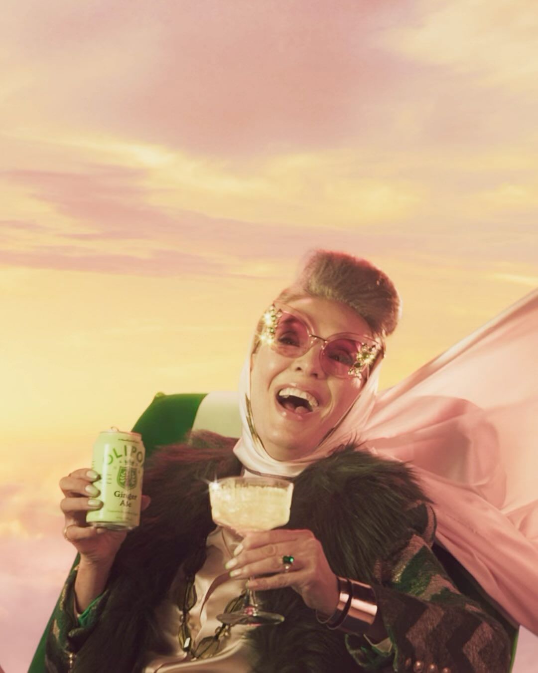 Cheerful woman with glasses raises a cocktail and canned drink in a pink sky, wearing a headscarf and fur-trimmed jacket.