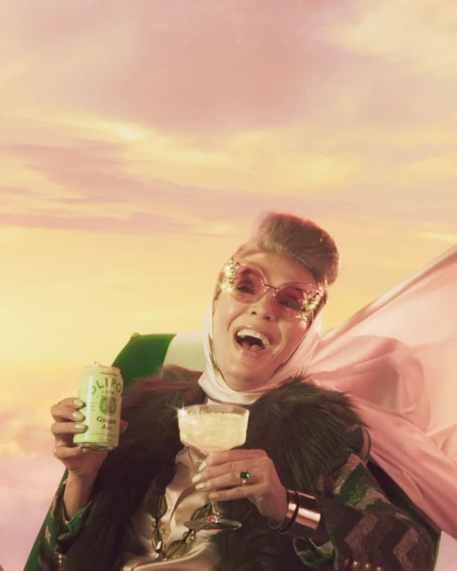 Cheerful woman with glasses raises a cocktail and canned drink in a pink sky, wearing a headscarf and fur-trimmed jacket.