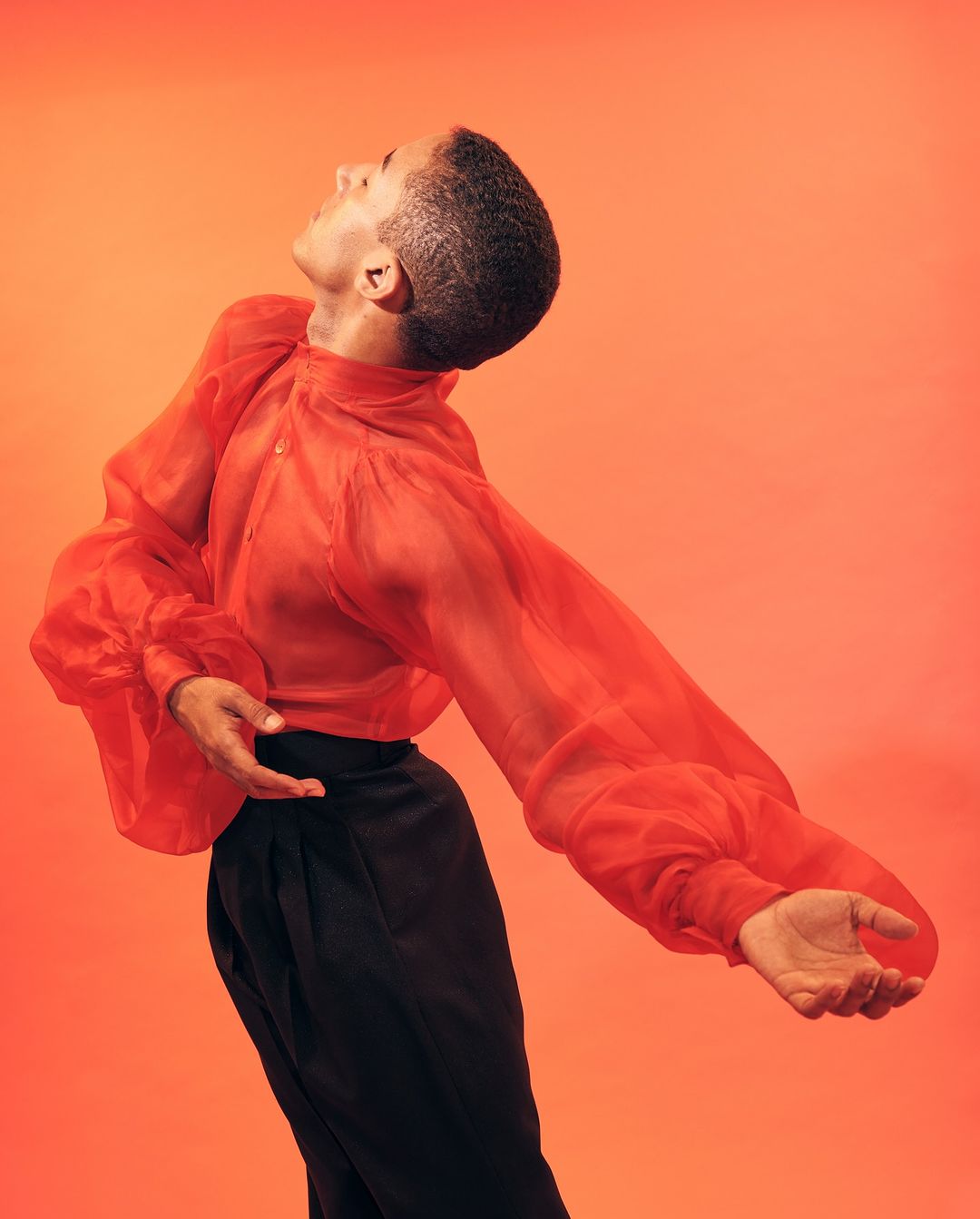 Side view of a Black adult in a sheer, orange-red blouse and black pants, leaning backward in a pose on a matching backdrop.