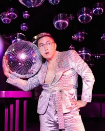 Man wearing a light patterned suit holding a disco ball near his head in a nightclub filled with hanging disco balls.