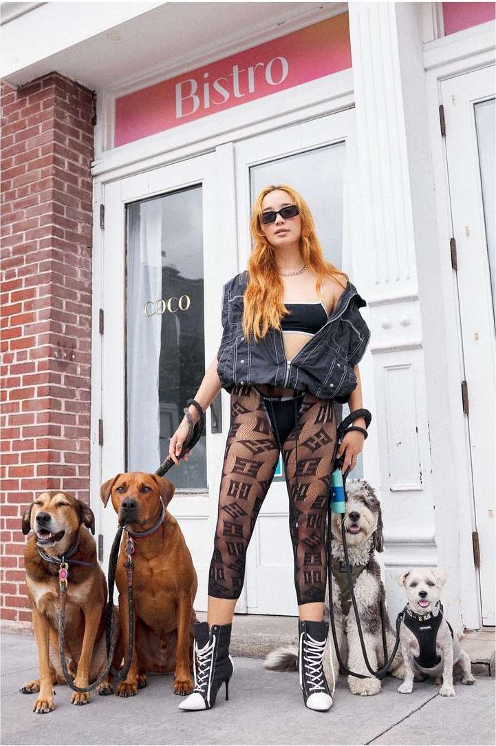 A woman in streetwear stands on a sidewalk with four dogs near a bistro entrance, showcasing an urban fashion style.