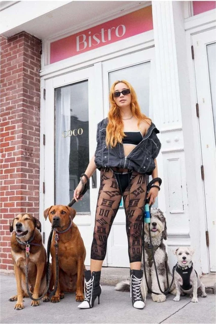 A woman in streetwear stands on a sidewalk with four dogs near a bistro entrance, showcasing an urban fashion style.