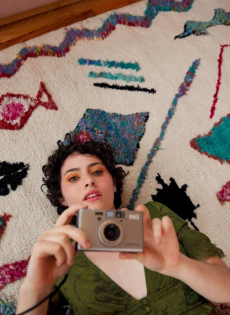 Young woman with curly hair lies on a colorful rug, holding a compact digital camera, creating a self-portrait in an indoor setting.