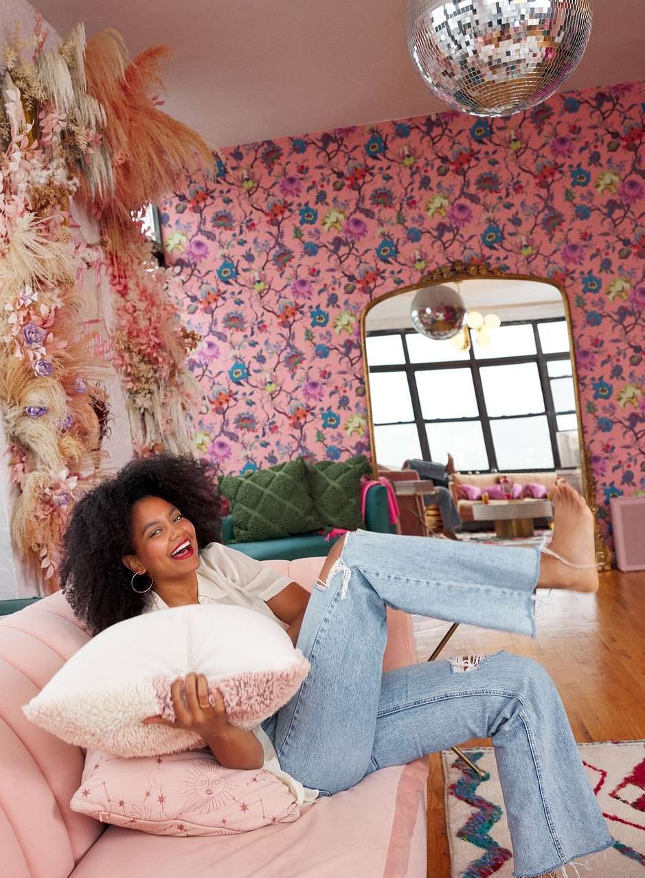 A woman relaxes on a pink couch in a pink floral living room. She smiles holding a pillow with a disco ball hanging overhead.