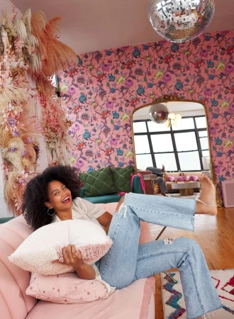 A woman relaxes on a pink couch in a pink floral living room. She smiles holding a pillow with a disco ball hanging overhead.