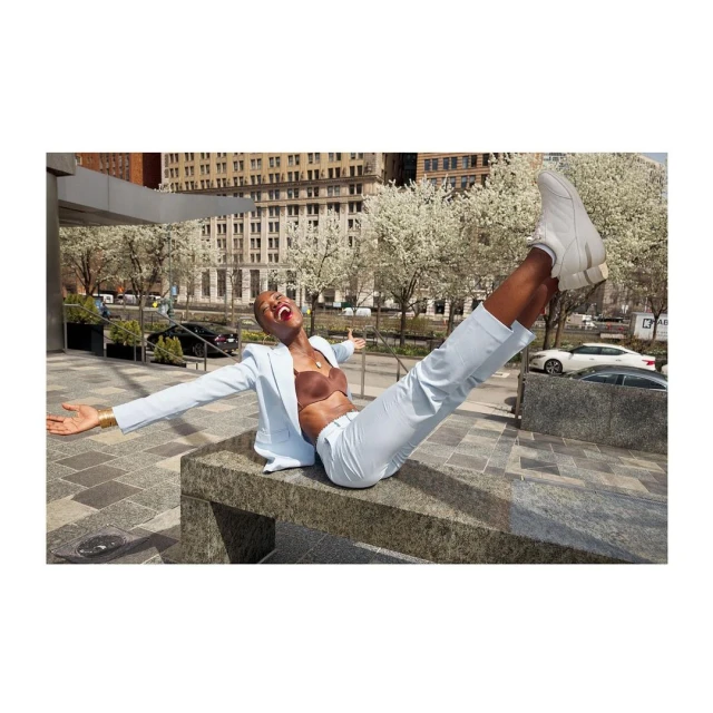 Woman in light blue suit and white sneakers poses outdoors on a stone bench in an urban setting with an ecstatic expression.
