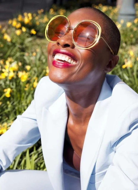 Smiling adult female wearing round sunglasses and a light blue suit jacket, set against a blurred background of yellow flowers.