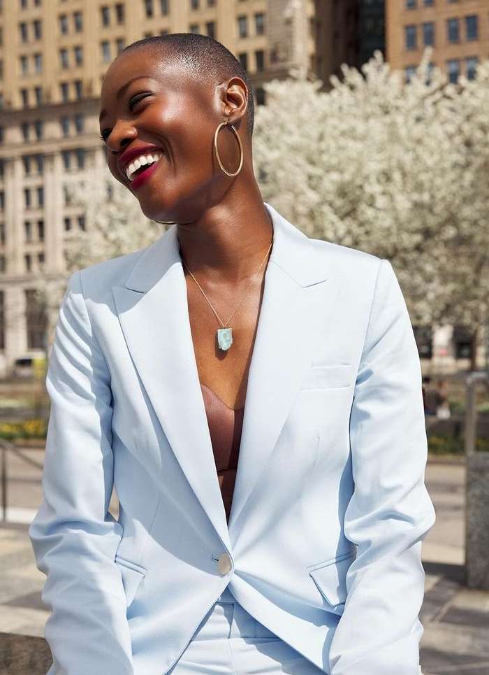 Smiling Black woman in light blue pantsuit, statement jewelry, posing outdoors in city, blooming trees, neutral background, and natural light.