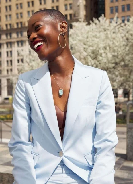 Smiling Black woman in light blue pantsuit, statement jewelry, posing outdoors in city, blooming trees, neutral background, and natural light.