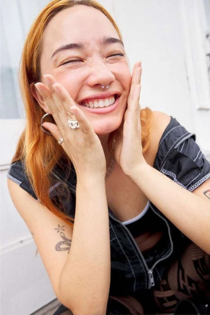 Smiling young woman with auburn hair and a septum piercing cupping her face with her hands in an outdoor setting.