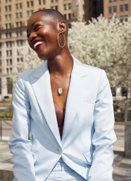 Smiling Black woman with a shaved head wearing a light blue blazer and large gold hoop earrings outdoors in New York.
