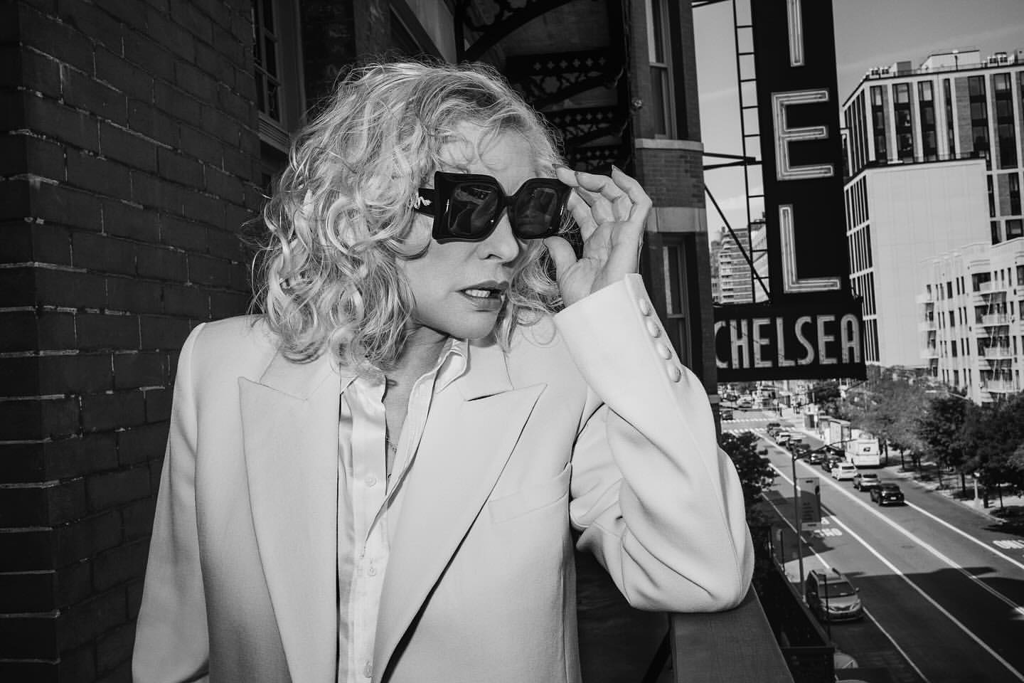 Black and white portrait of a stylish adult female with curly blonde hair, large sunglasses, and a light-colored blazer on a balcony.