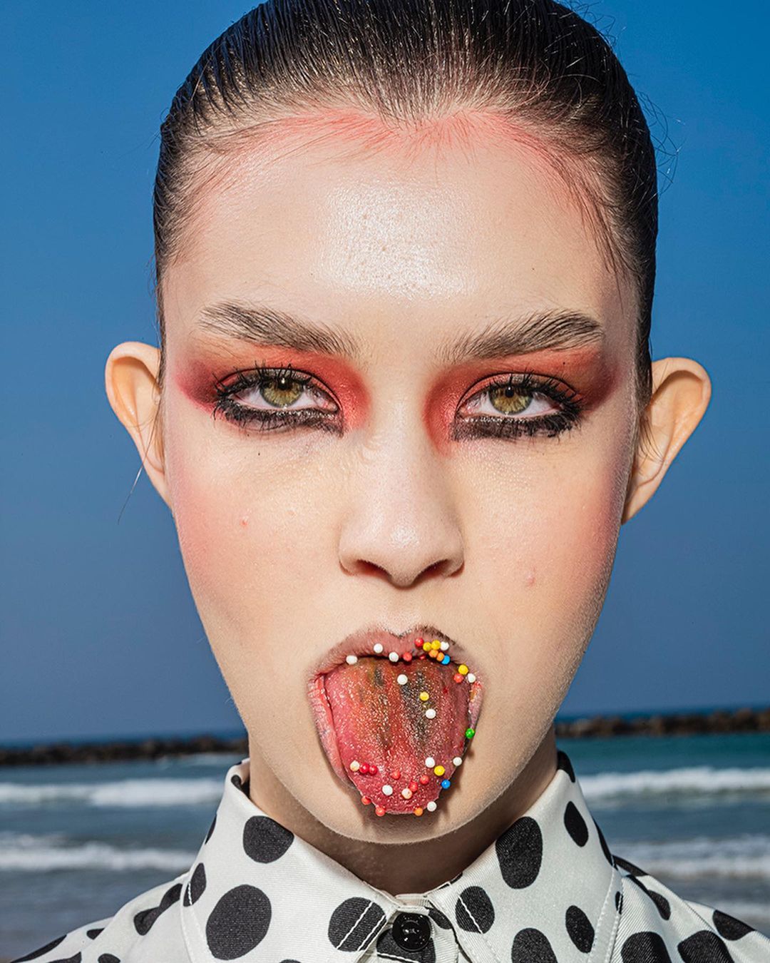 Portrait of a woman with colorful makeup, tongue out, and sprinkles on her tongue, set against a beach and blue sky backdrop.