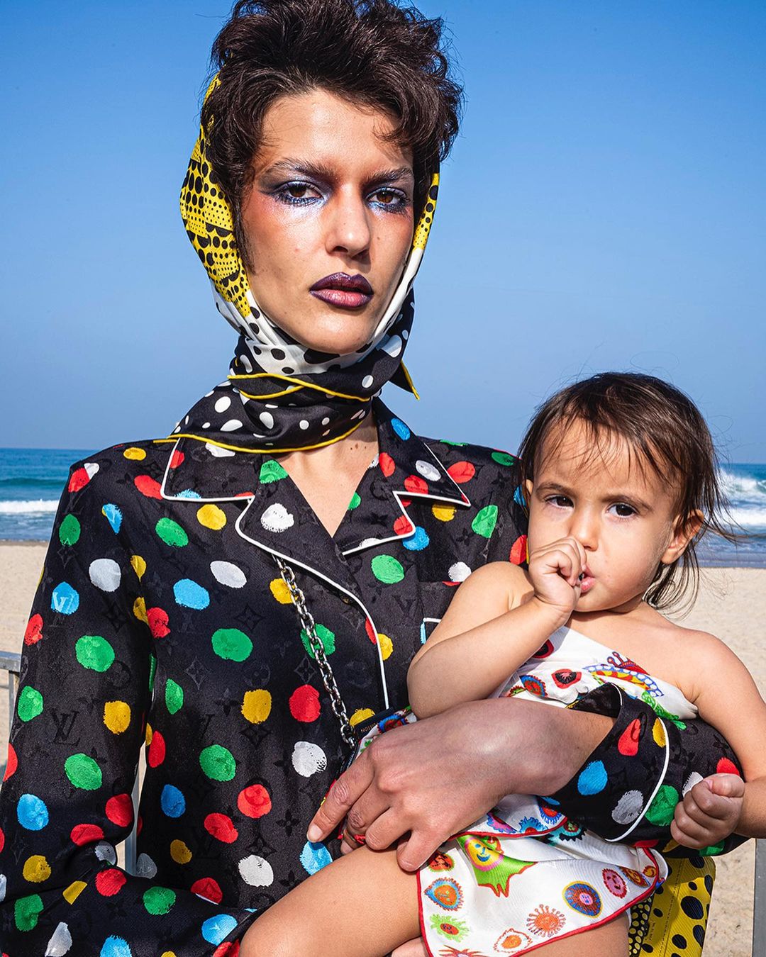 A woman in a patterned shirt and headscarf holds a baby on a beach with blue sky and ocean in the background during daytime.