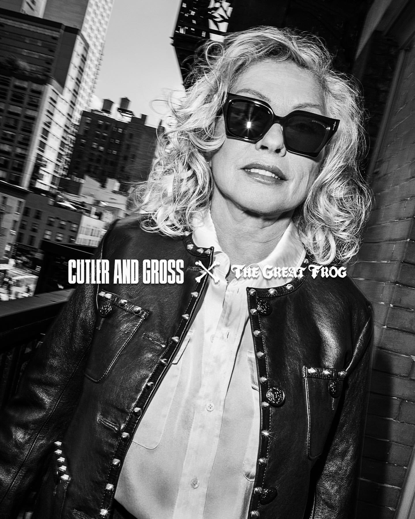 Black and white portrait of a woman with curly hair wearing sunglasses and a leather jacket in a city environment with buildings.