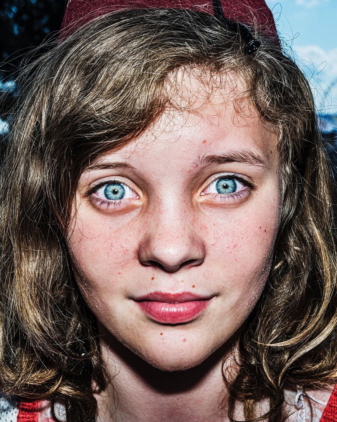 Close-up portrait of a young female with light skin, curly blonde hair and blue eyes wearing a maroon hat with a serious expression.