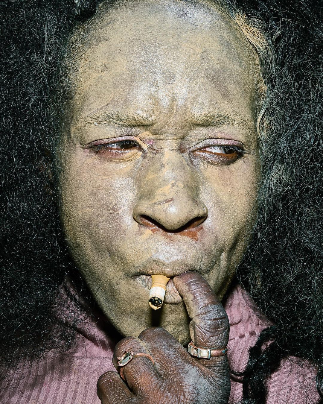 Close-up portrait of an adult woman with olive-toned skin, a cigarette in her mouth, wearing a purple top with vertical stripes indoors.