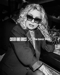 Black and white portrait of Debbie Harry wearing dark oversized sunglasses and a dark blazer in a dimly lit bar setting.