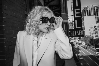 Blonde woman wearing oversized sunglasses and a tailored blazer on a balcony overlooking a city street.