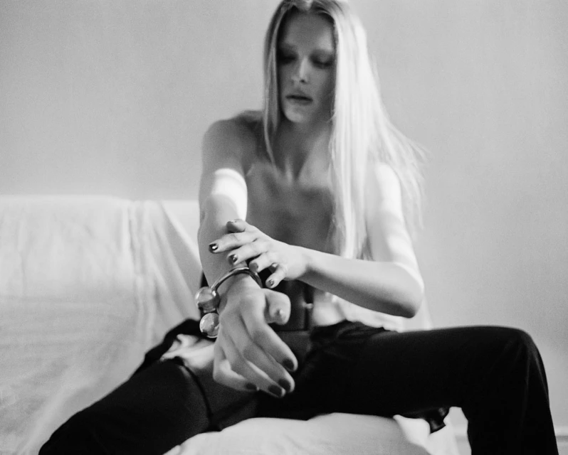 A young adult woman with long blonde hair sits in a brightly lit room wearing dark pants and a metal bracelet. Black and white image.